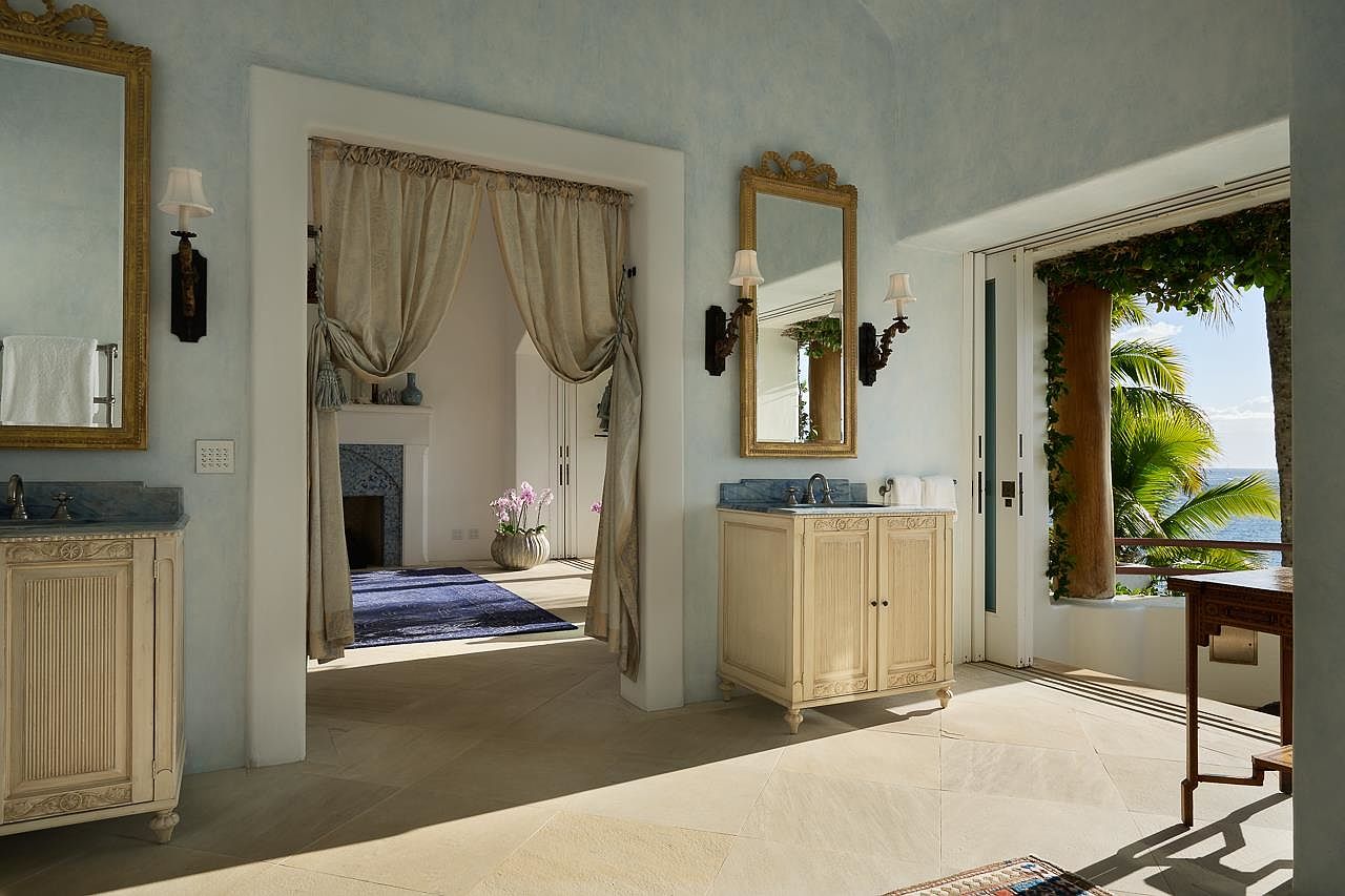 This is a luxurious primary bathroom featuring dual vanities with marble countertops and ornate mirrors. A doorway with elegant curtains leads to another room, while a door opens to a balcony with ocean views. The bathroom has a serene and sophisticated ambiance, perfect for relaxation.