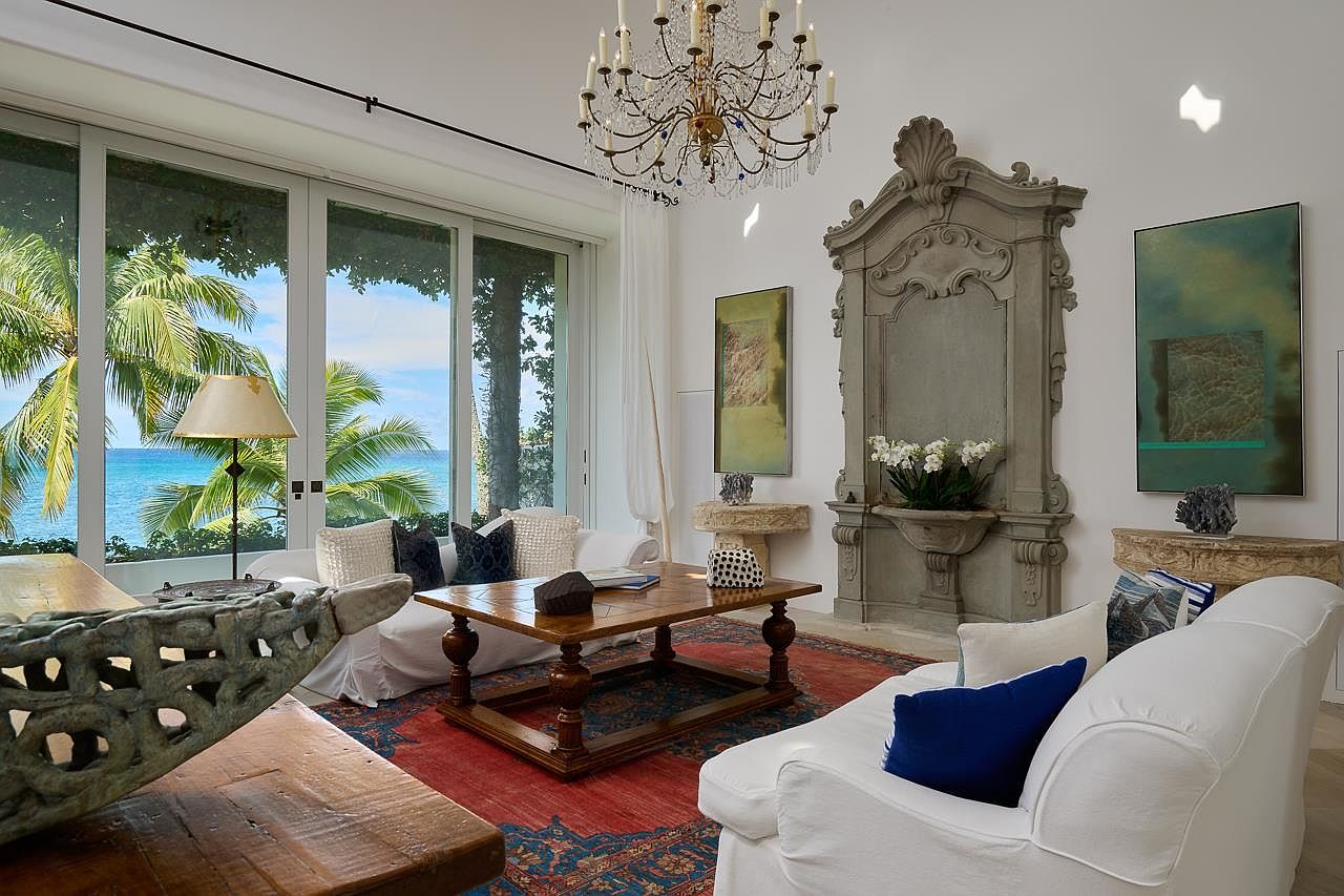 This is an interior shot of a luxurious living room featuring a blend of classic and coastal styles. The room is bright and airy, with large windows offering ocean views framed by palm trees. Key elements include a decorative chandelier, ornate architectural details, comfortable white sofas, and a rich red and blue patterned rug, creating an inviting and elegant space.