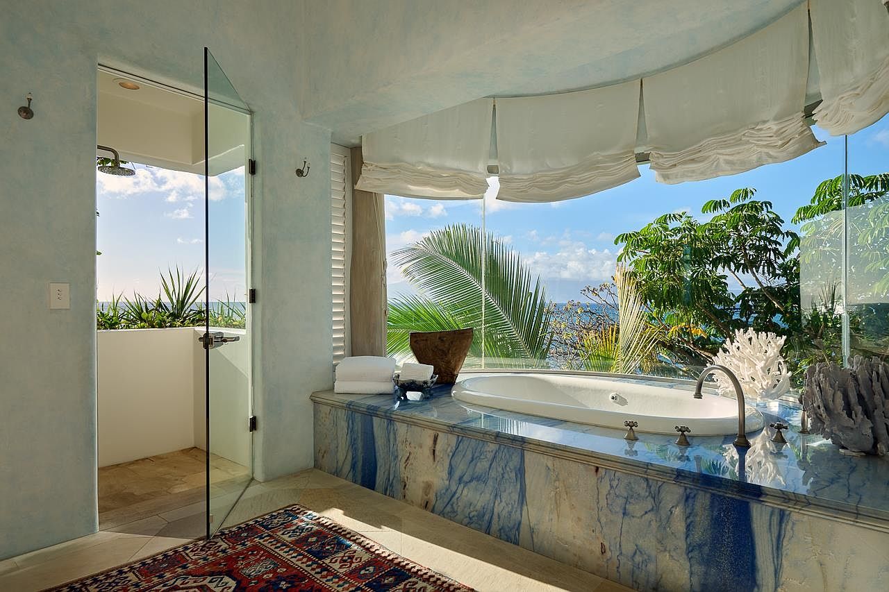 This is a luxurious primary bathroom featuring a large soaking tub with a blue marble surround, offering stunning ocean views through a wide window. The bathroom includes a glass-enclosed shower and is decorated with natural elements like coral, creating a spa-like atmosphere. A patterned rug adds warmth to the light-colored flooring.