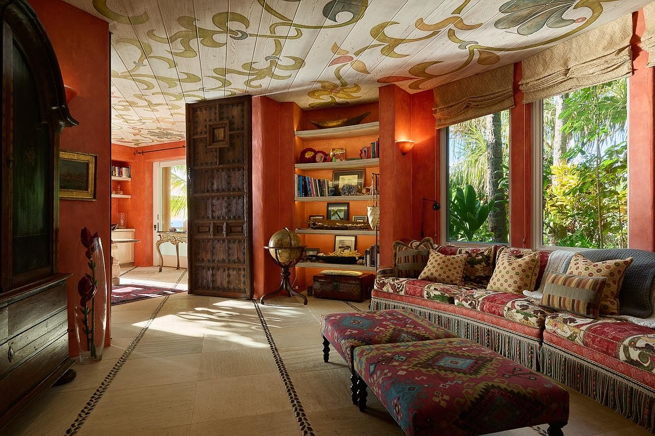 This interior shot showcases a vibrant living room with a unique, hand-painted ceiling design. The room features a comfortable sofa adorned with patterned cushions and a fringed edge, complemented by matching ottomans. A large wooden door and built-in bookshelves add character, while natural light streams in through the windows, creating a warm and inviting atmosphere.