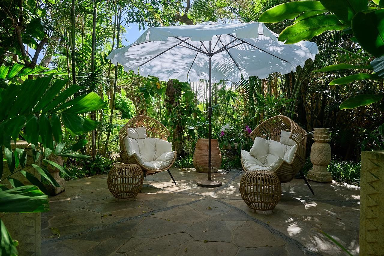 This image showcases a beautifully designed outdoor patio area, featuring two wicker chairs with plush white cushions and matching side tables, all sheltered by a large white umbrella. The patio is surrounded by lush tropical foliage, creating a serene and private retreat. The stone flooring adds a touch of rustic elegance to the space.