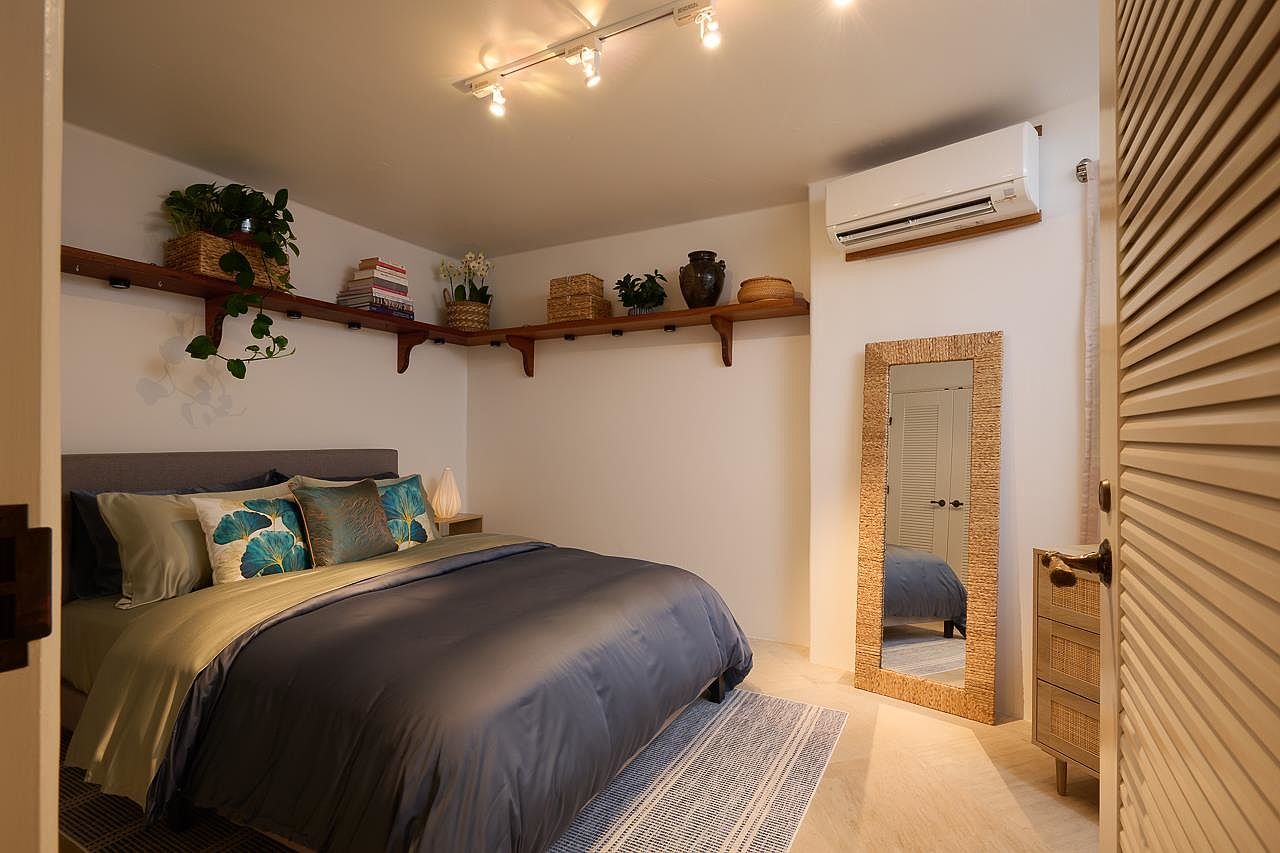 This is a cozy bedroom featuring a queen-sized bed with a gray headboard and blue and green bedding. The room has a warm and inviting atmosphere, enhanced by the natural light and the wooden shelves displaying plants and decorative items. A full-length mirror and a small dresser add functionality to the space.