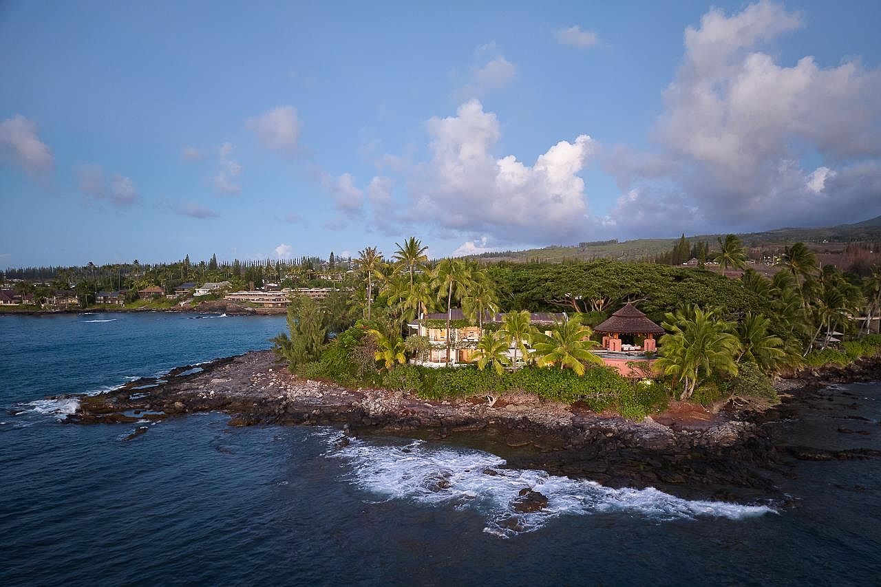 This aerial view showcases a luxurious property nestled on a rocky coastline, surrounded by lush tropical vegetation and palm trees. The residence features a charming gazebo and appears to offer stunning ocean views. The scene is set against a backdrop of a partly cloudy sky and distant hills, creating an idyllic and exclusive atmosphere.