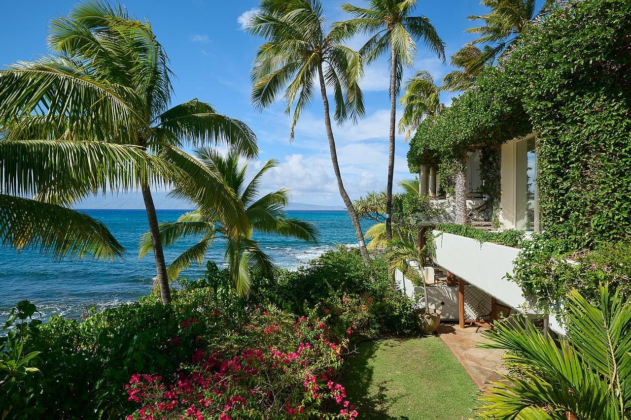 This image showcases a lush tropical yard with vibrant greenery and pink bougainvillea flowers, overlooking a serene ocean view. Palm trees frame the scene, adding to the tropical ambiance, while a portion of a building covered in climbing plants is visible on the right, suggesting a well-maintained and luxurious property. The overall impression is one of tranquility and natural beauty, ideal for a relaxing coastal lifestyle.
