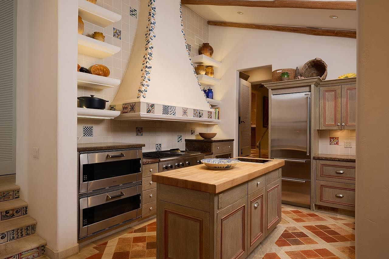 This is a well-appointed kitchen featuring custom cabinetry with a distressed finish, stainless steel appliances, and a butcher block island. The kitchen has a rustic yet elegant feel, with decorative tile accents and a unique range hood design. The flooring is patterned tile, adding to the overall charm of the space.