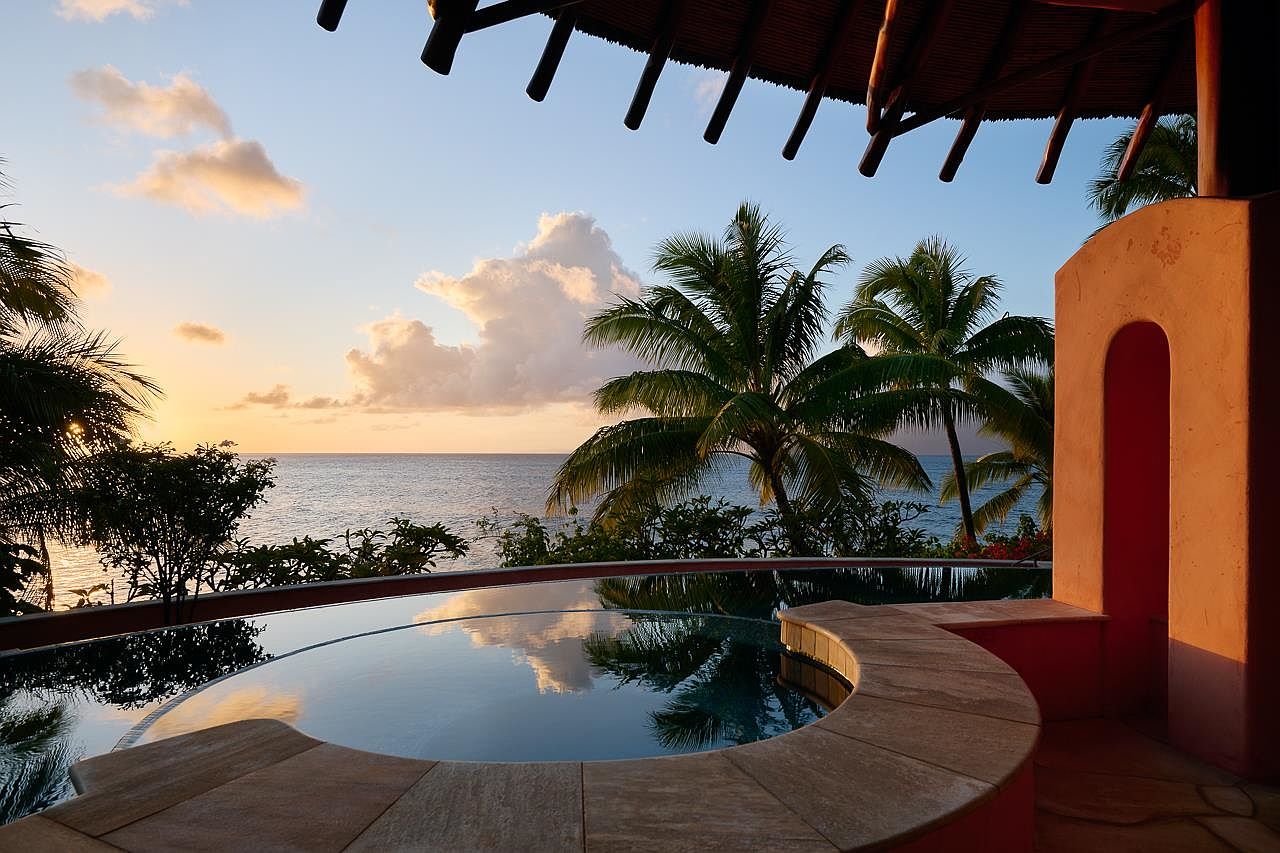 This image showcases a luxurious outdoor pool and spa area with a stunning ocean view at sunset. The curved pool is surrounded by stone tiling and lush tropical vegetation, including palm trees. An arched architectural feature adds a touch of elegance, while the overall scene evokes a sense of tranquility and relaxation, perfect for a high-end real estate listing.