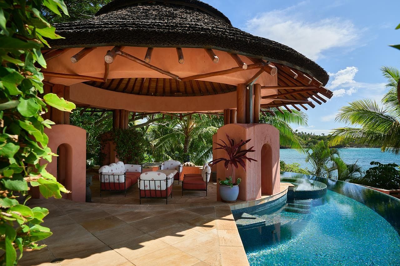 This image showcases a luxurious outdoor space featuring a unique pool design with integrated steps and a curved edge overlooking a serene body of water. A thatched-roof gazebo with comfortable seating provides a shaded lounge area, complemented by lush tropical vegetation. The overall impression is one of relaxation and high-end resort living.