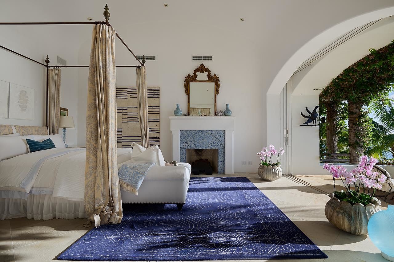 This is a luxurious primary bedroom featuring a four-poster bed with elegant drapery, a white sofa, and a striking blue patterned rug. A fireplace with a decorative mirror above it adds a focal point, while an arched doorway leads to an outdoor space with lush greenery. The room exudes a sense of calm and sophistication.