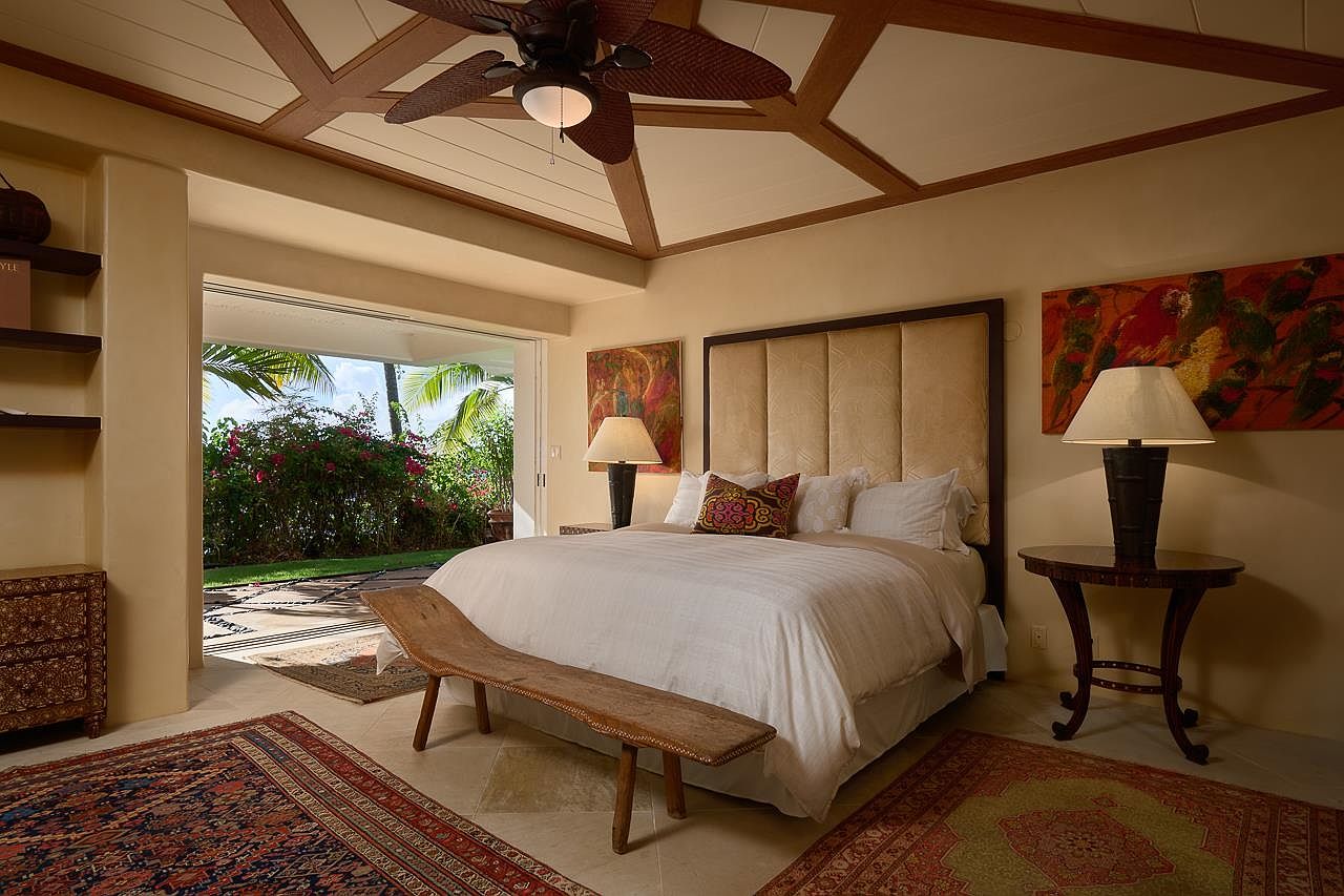 This is a primary bedroom featuring a king-size bed with a padded headboard, flanked by two nightstands with lamps. The room has a tropical feel, with an open doorway leading to a lush garden. The ceiling has a unique design with exposed beams and a ceiling fan, and the room is decorated with colorful artwork and oriental rugs.