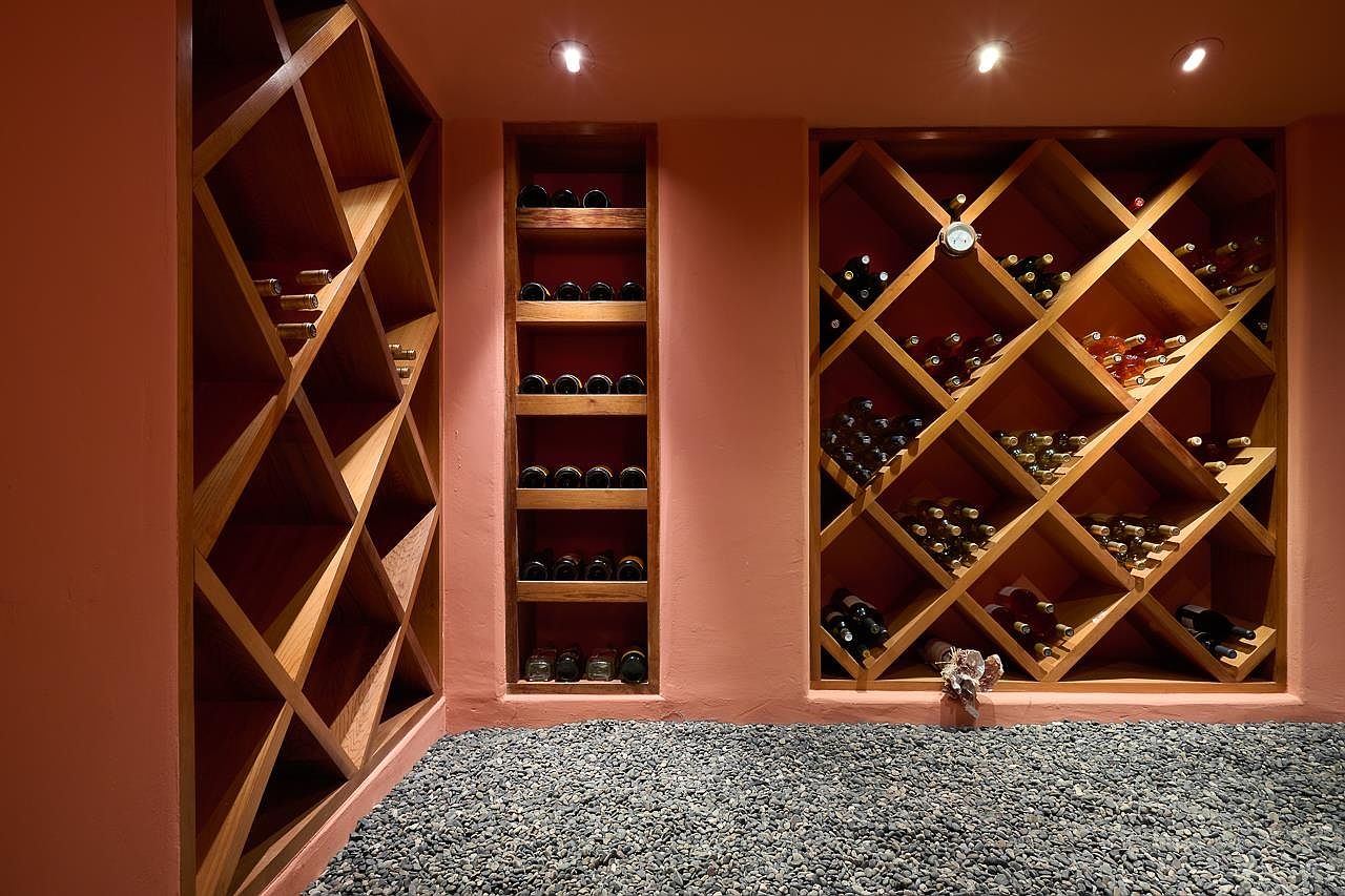 This wine cellar features custom wooden wine racks built into the walls, showcasing a variety of wine bottles. The walls are painted in a warm, earthy tone, and the floor is covered with small, dark pebbles, adding a unique textural element. Recessed lighting highlights the wine collection, creating an inviting and sophisticated atmosphere.