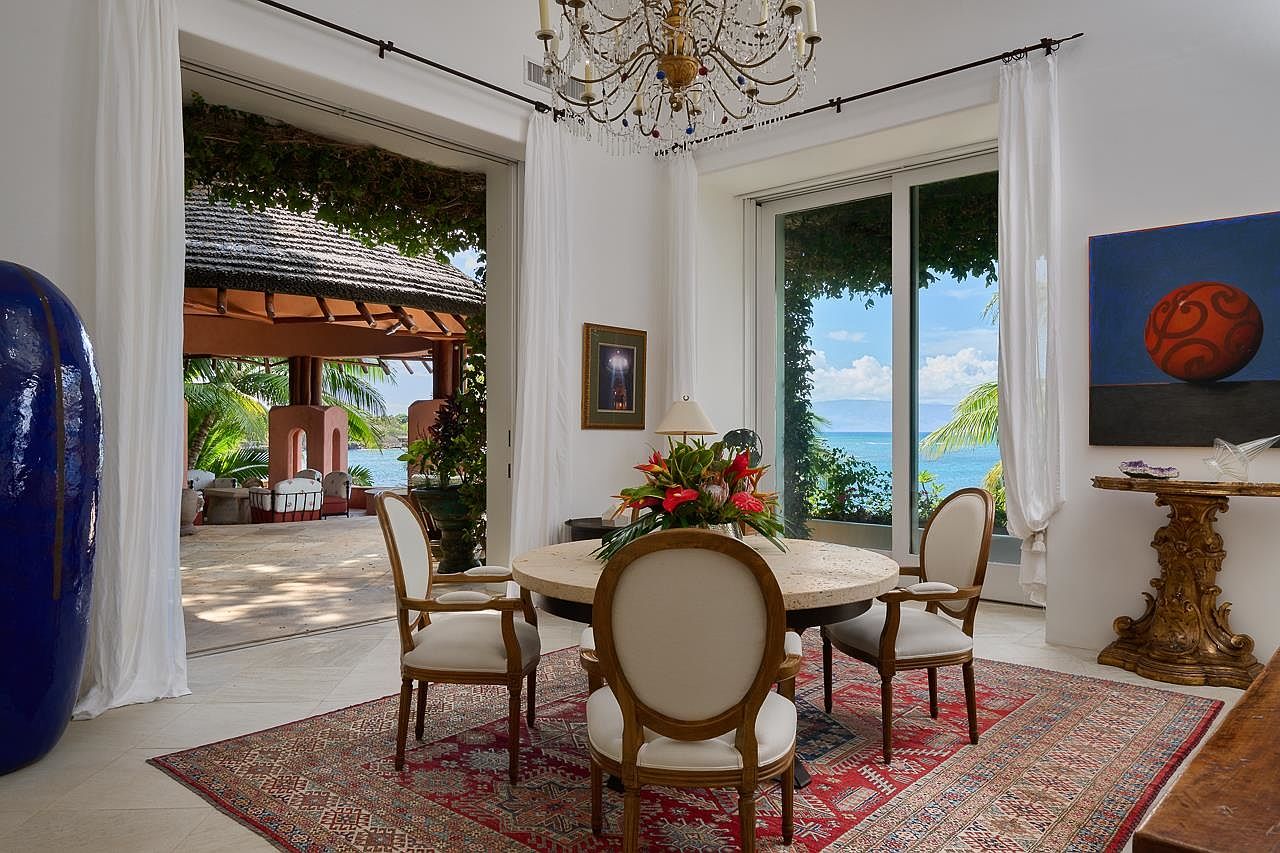 This is an elegant dining room featuring a round table with four chairs, a chandelier, and a large rug. The room opens to an outdoor patio with a gazebo and ocean view, creating a seamless indoor-outdoor living experience. A painting and decorative table add to the room's sophisticated ambiance.