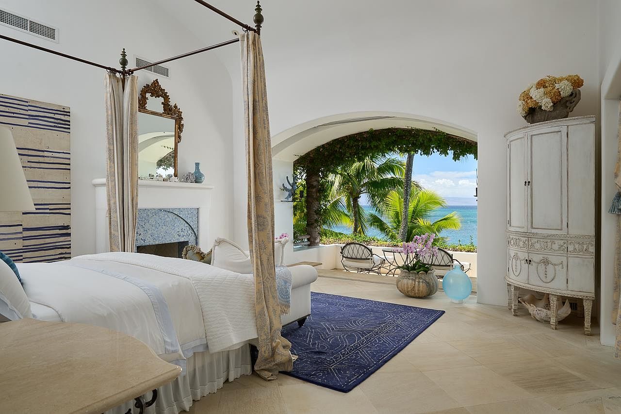 This primary bedroom features a four-poster bed with elegant drapery, a decorative fireplace, and a stunning ocean view through an arched opening. The room is decorated in a light, airy style with a blue accent rug and a vintage-style armoire. The overall impression is luxurious and serene, emphasizing the connection to the beautiful outdoor scenery.