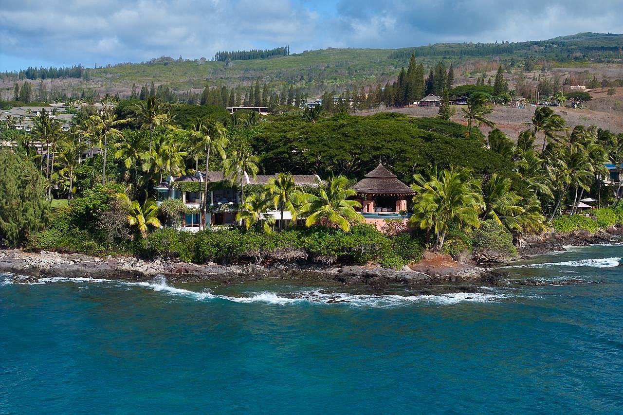 This aerial view showcases a luxurious beachfront property nestled among lush tropical vegetation. The estate features multiple buildings, including a distinctive round structure with a thatched roof, all situated along a rocky coastline with turquoise waters. The overall impression is one of secluded elegance and tropical paradise.