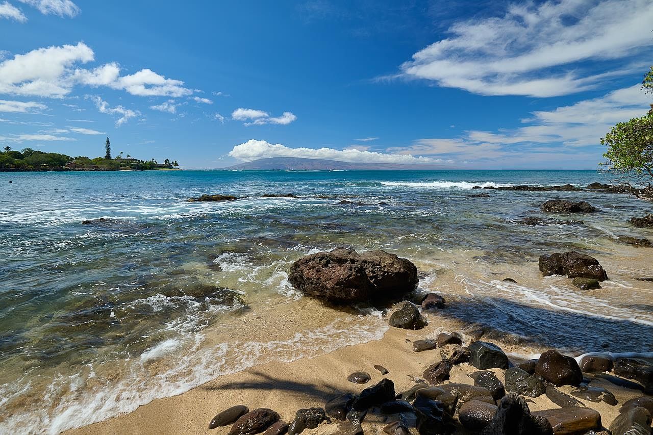 This picturesque beach scene showcases a tranquil yard/garden area with a sandy beach and clear turquoise water. Large rocks are scattered along the shoreline, adding texture and visual interest. The view extends to a distant island under a bright blue sky with scattered clouds, creating a serene and inviting atmosphere.