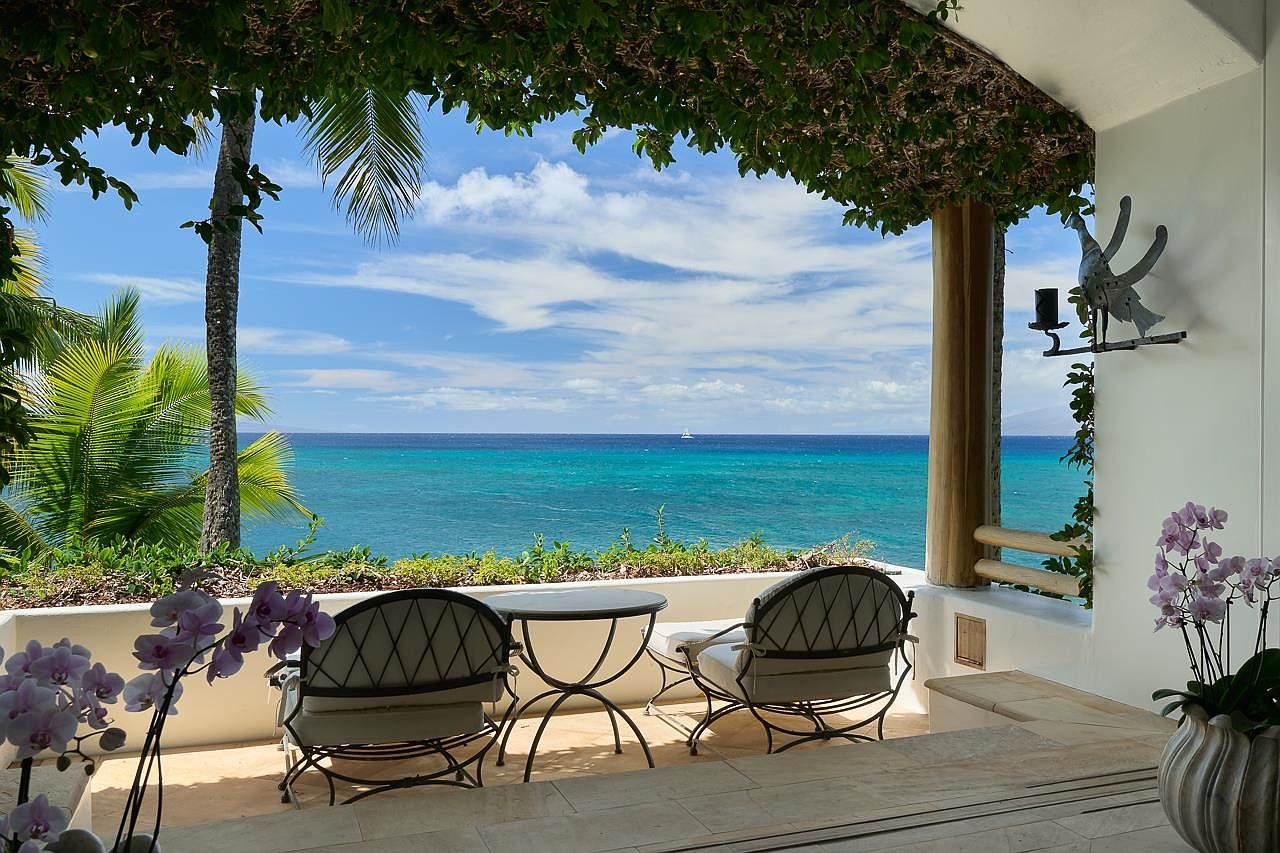 This image showcases a serene patio or balcony with a stunning ocean view. Two wrought-iron chairs and a small table are arranged on the tiled surface, inviting relaxation. Lush greenery frames the scene, creating a tranquil and luxurious outdoor living space perfect for enjoying the coastal scenery.