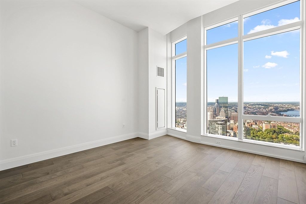 This bright and minimalist bedroom features expansive floor-to-ceiling windows that offer a breathtaking, high-altitude panoramic view of the city skyline and surrounding greenery. The room is finished with clean white walls and warm-toned wood-look flooring, creating a serene and modern atmosphere. The perspective is captured from the corner of the room, emphasizing the open, airy feel and the impressive architectural integration of the urban landscape.