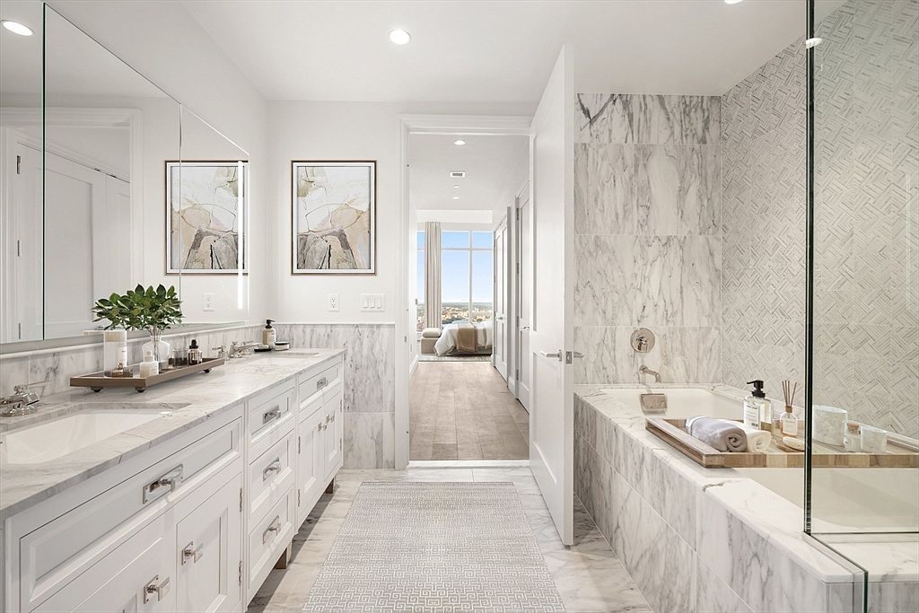 This luxurious primary bathroom features elegant marble surfaces throughout, including a double vanity and a deep soaking tub integrated into a marble-clad surround. A glass-enclosed shower area and a patterned tile accent wall add sophisticated texture, while a doorway provides a seamless transition into a bright, airy bedroom beyond. The space exudes a high-end, spa-like atmosphere with clean lines and a bright, neutral color palette.