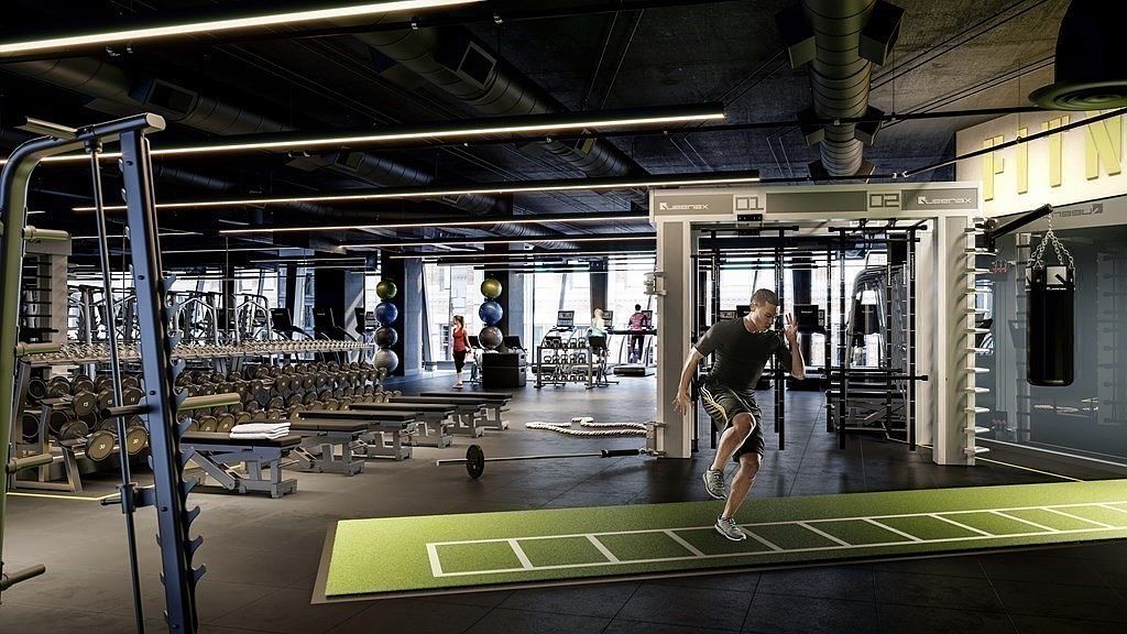 This expansive, modern fitness center features a high-end, industrial aesthetic with exposed ceilings, sleek black lighting fixtures, and a dedicated agility training area with a green turf ladder. The space is well-equipped with a variety of professional-grade strength training machines, a rack of dumbbells, and a functional training rig, creating an inviting atmosphere for a comprehensive workout. The perspective is a wide-angle shot from the floor level, capturing the depth of the facility and highlighting the active, energetic environment.