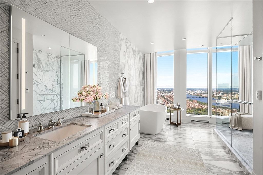 This luxurious primary bathroom features a sophisticated marble vanity with dual sinks, a freestanding soaking tub, and a spacious glass-enclosed walk-in shower. Floor-to-ceiling windows offer a breathtaking, panoramic view of the city skyline and river, bathing the room in natural light. The elegant design is complemented by intricate tile work and high-end finishes, creating a serene and opulent spa-like atmosphere.