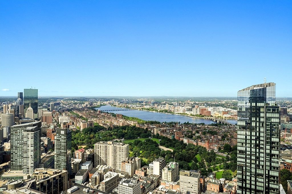 This high-altitude aerial view captures a sprawling urban cityscape, prominently featuring a large river winding through the center and a lush green park nestled among dense residential and commercial buildings. The perspective highlights the impressive scale of the city, showcasing a mix of modern glass skyscrapers and historic architecture under a clear, expansive blue sky. It provides a comprehensive sense of location and urban density, emphasizing the property's prime position within the metropolitan landscape.