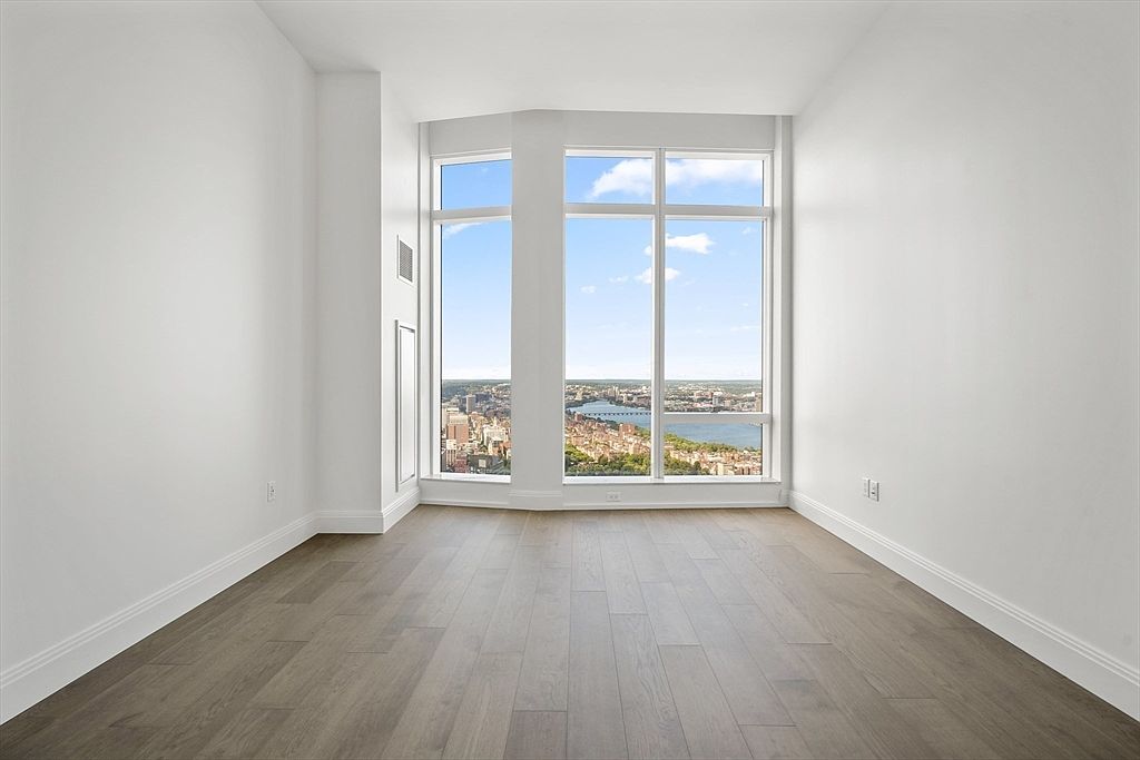 This bright and minimalist room features light-toned hardwood flooring and crisp white walls, creating a clean and airy atmosphere. The focal point is a large, floor-to-ceiling window assembly that offers a stunning, expansive panoramic view of a city skyline and a river. The perspective is centered and symmetrical, emphasizing the height and the impressive urban vista.