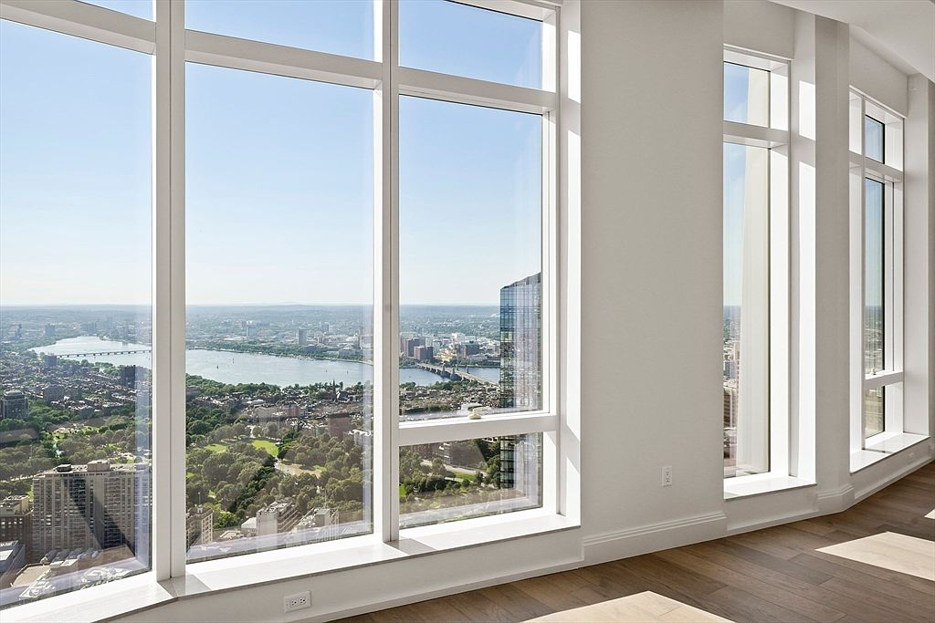 This high-rise interior space features floor-to-ceiling windows that offer a breathtaking, panoramic aerial view of a city skyline, a river, and a large park. The room is characterized by minimalist white walls, clean architectural lines, and warm-toned hardwood flooring, creating a bright and airy atmosphere. The perspective emphasizes the luxury of the height and the seamless connection between the modern living space and the expansive urban landscape outside.