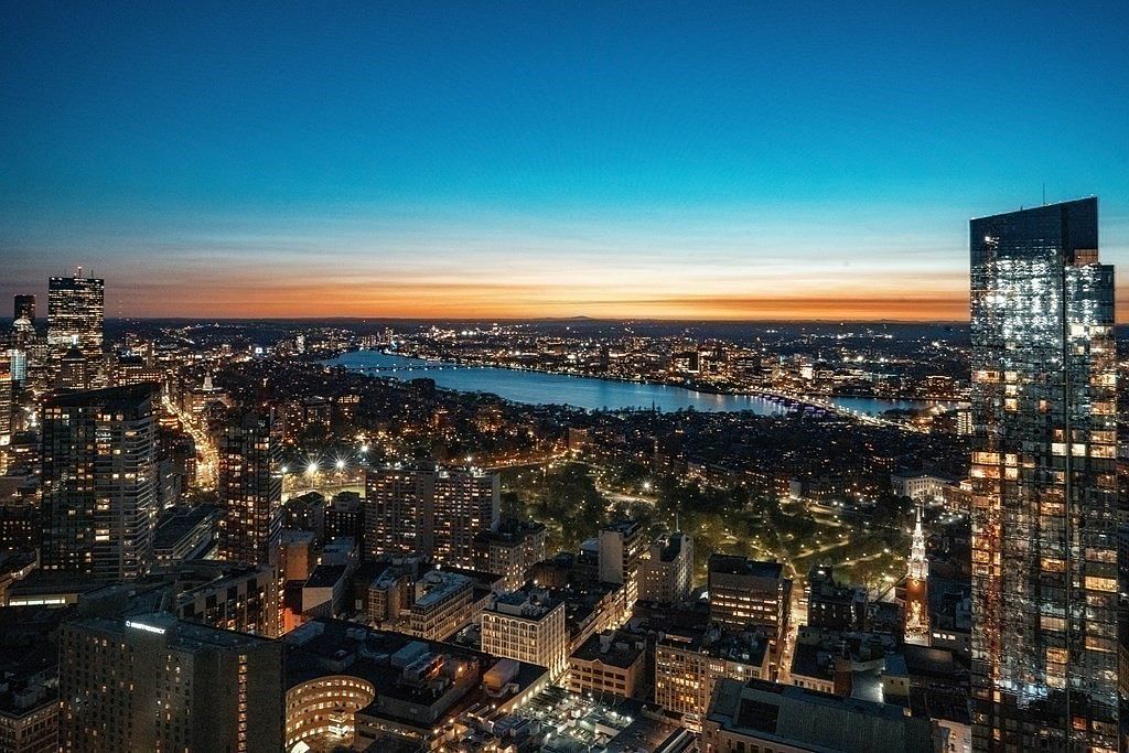 This stunning high-altitude aerial view captures a sprawling urban skyline at dusk, featuring a prominent modern glass skyscraper in the foreground and a serene river winding through the city. The horizon is painted with a vibrant sunset gradient, while the city below is illuminated by a dense network of streetlights and building lights. The perspective offers a cinematic, expansive look at the metropolitan landscape, highlighting the prime location and architectural density of the area.
