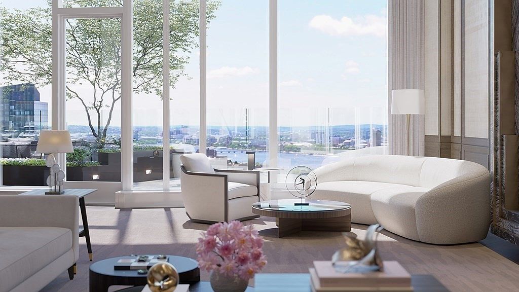 This sophisticated, high-end living room features a curved, cream-colored sofa and a modern armchair, centered around a circular glass-topped coffee table. Floor-to-ceiling windows provide an expansive, panoramic view of a city skyline and river, bathing the room in natural light. The interior design is elegant and contemporary, characterized by neutral tones, plush textures, and refined decorative accents.