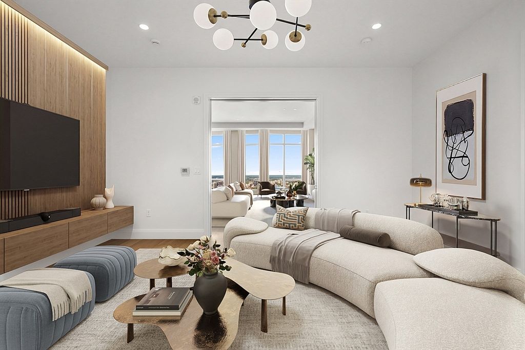 This sophisticated living room features a contemporary curved cream sofa, a pair of blue upholstered ottomans, and a multi-tiered organic-shaped coffee table. The space is anchored by a sleek wood-paneled media wall with a built-in floating console and a modern globe chandelier overhead. Through the central doorway, a secondary living area with floor-to-ceiling windows offers a stunning, expansive view of the city skyline.