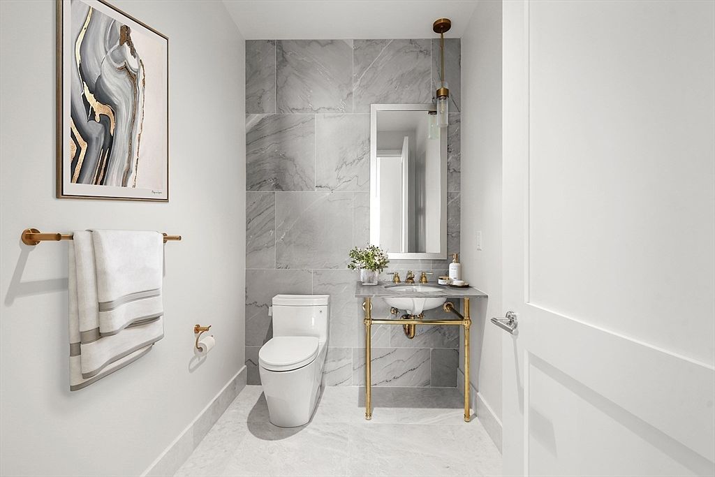 This elegant powder room features a sophisticated marble-tiled accent wall and matching flooring, creating a cohesive and luxurious aesthetic. The space is anchored by a unique pedestal sink with gold-toned plumbing fixtures, complemented by a sleek mirror and modern wall sconce. A framed abstract art piece adds a touch of contemporary flair, while the overall design feels bright, clean, and high-end.