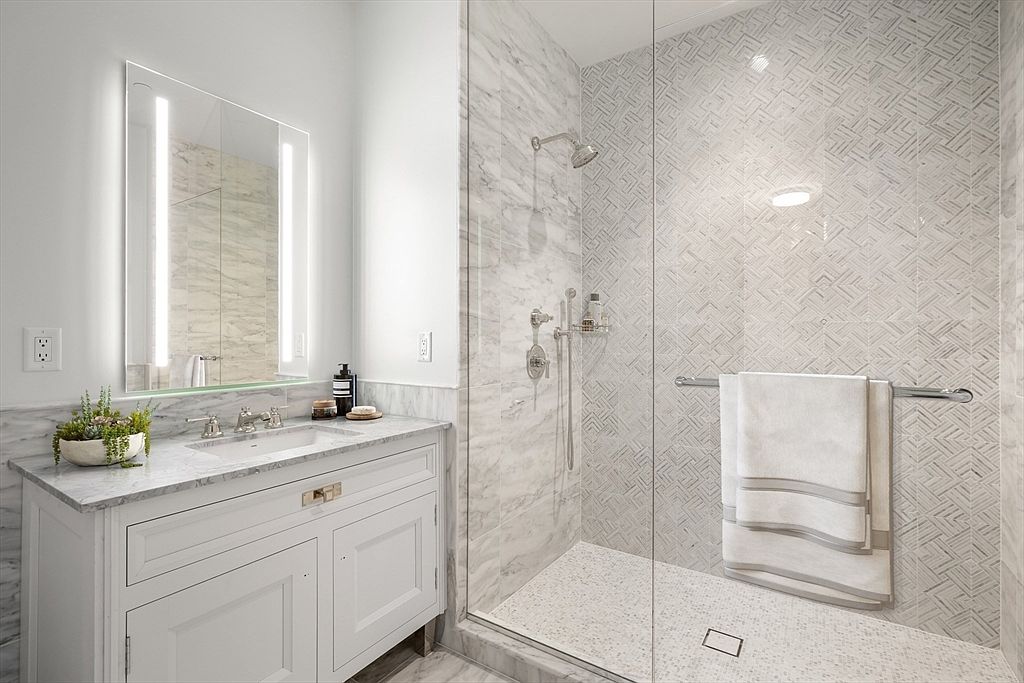 This elegant primary bathroom features a sleek white vanity with a marble countertop and a modern, backlit rectangular mirror. To the right, a spacious walk-in shower is enclosed by a glass panel, showcasing intricate patterned tile work on the back wall and a clean, minimalist design. The overall aesthetic is bright, luxurious, and contemporary, emphasizing high-end finishes and a spa-like atmosphere.