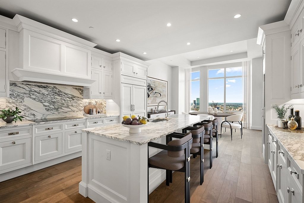 This sophisticated, high-end kitchen features crisp white cabinetry, a striking marble-veined backsplash, and a large central island topped with matching stone. The space is bathed in natural light from floor-to-ceiling windows that offer a panoramic city view, complemented by warm hardwood flooring and elegant bar seating. The perspective is a wide-angle shot from the kitchen entrance, emphasizing the open-concept layout and luxurious, modern aesthetic.