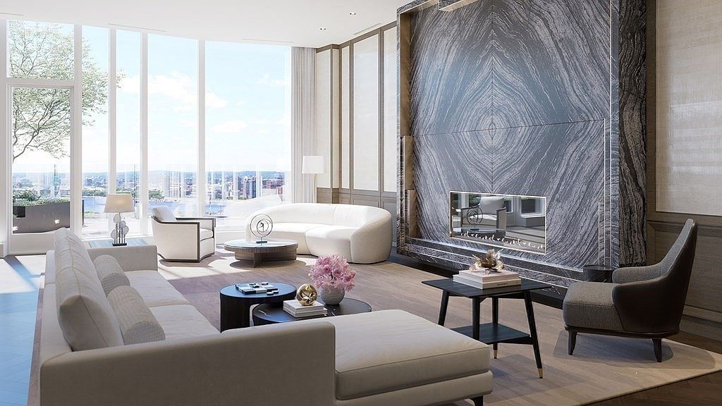 This sophisticated, high-end living room features a striking floor-to-ceiling marble fireplace as its focal point, complemented by plush, contemporary cream-colored seating. Large, expansive windows flood the space with natural light while offering a panoramic view of the city skyline and waterfront. The room is elegantly furnished with layered coffee tables, a textured area rug, and tasteful decor, creating an atmosphere of refined luxury and comfort.