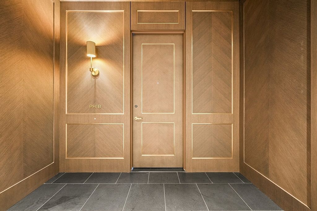 This elegant entryway features sophisticated wood paneling with a chevron pattern and gold trim, creating a luxurious and warm atmosphere. A modern wall sconce provides soft, ambient lighting, while the dark tiled floor offers a sharp, clean contrast to the rich wood tones. The centered door, marked 'PH1B', serves as a focal point, suggesting a high-end residential unit.