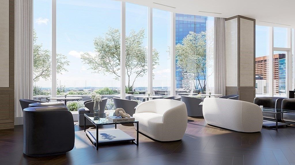 This sophisticated lounge area features a modern, minimalist aesthetic with plush, curved white sofas and dark leather armchairs arranged on a rich, dark herringbone wood floor. Floor-to-ceiling windows flood the space with natural light, offering a panoramic view of an outdoor terrace and the surrounding cityscape. The elegant decor and open layout create a luxurious, inviting atmosphere perfect for social gatherings or quiet relaxation.