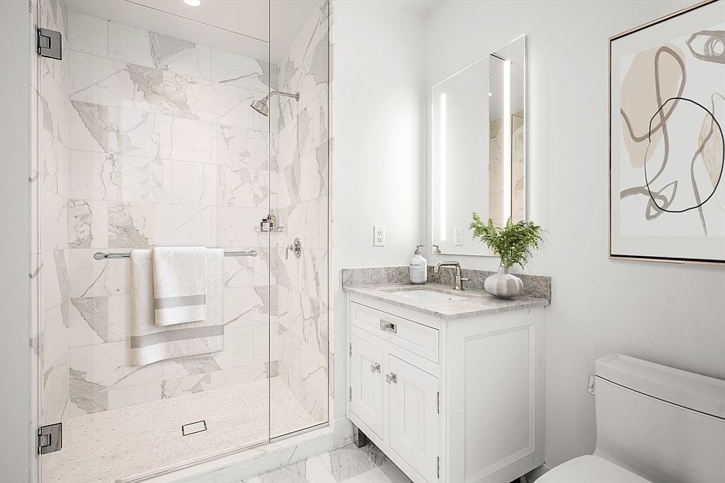 This bright and modern bathroom features a sleek glass-enclosed walk-in shower with elegant marble-patterned tile walls and a textured floor. A white vanity with a stone countertop and a contemporary mirror with integrated lighting creates a clean, sophisticated aesthetic. The space is accented by a minimalist framed piece of art on the wall, contributing to a polished and inviting atmosphere.