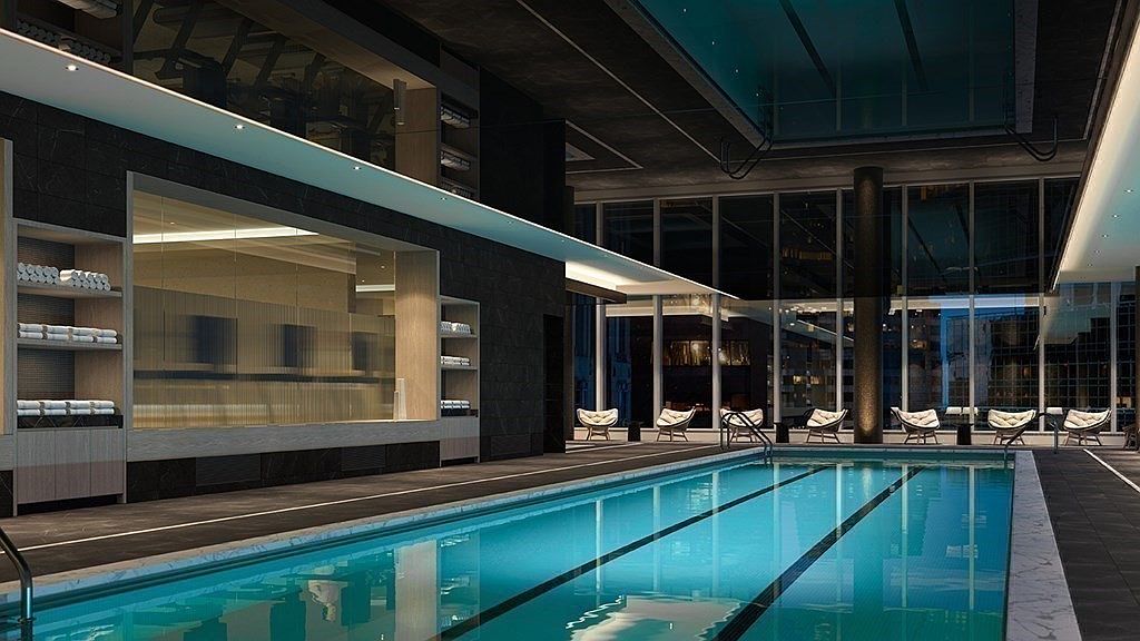 This sophisticated indoor lap pool area exudes luxury with its sleek, modern design and floor-to-ceiling windows overlooking a city skyline. The space features dark stone flooring, minimalist lounge seating, and integrated shelving with neatly stacked towels, creating a high-end spa-like atmosphere. The perspective is a wide, eye-level shot that emphasizes the length of the pool and the seamless transition between the interior amenities and the urban environment outside.