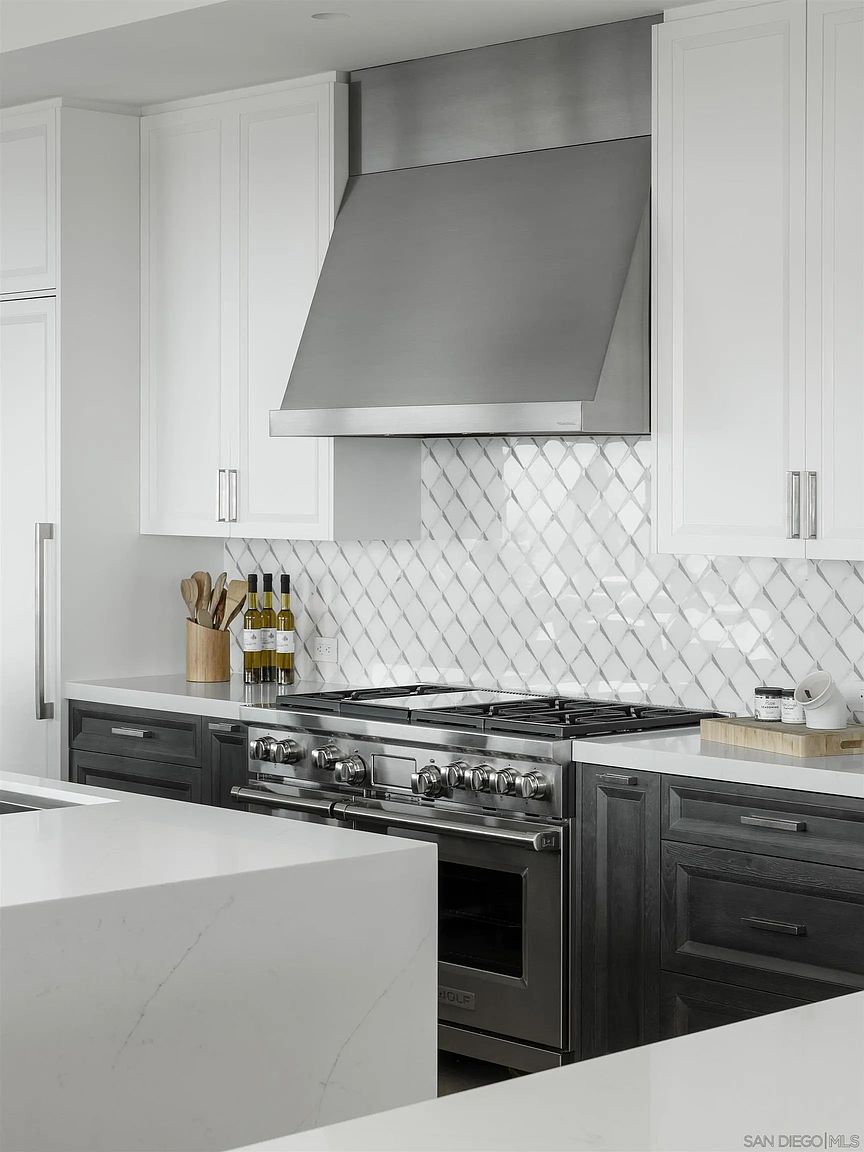 This is a well-lit kitchen featuring a stainless steel range hood above a professional-grade stove. The cabinetry is a mix of white and dark wood, complemented by white countertops and a textured white backsplash. The kitchen island is visible in the foreground, suggesting a spacious and modern design.