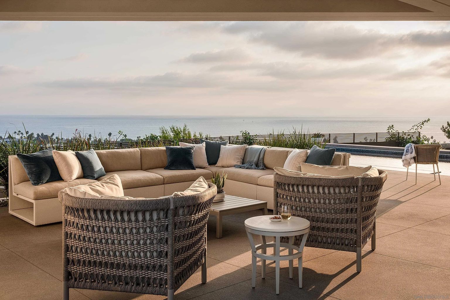 This image showcases a luxurious outdoor patio or deck area, featuring comfortable sectional seating with plush pillows, complemented by stylish wicker chairs and a small side table. The space overlooks a stunning ocean view, creating a serene and inviting atmosphere perfect for relaxation and entertaining. The overall design emphasizes comfort and elegance, making it an ideal selling point for a high-end property.