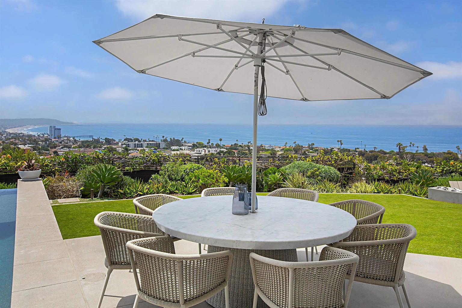 This image showcases an elegant outdoor patio area, perfect for entertaining or relaxing. A round marble table with woven chairs sits under a large white umbrella, offering shade and style. The patio overlooks a lush green lawn and a stunning ocean view, creating a serene and luxurious atmosphere.