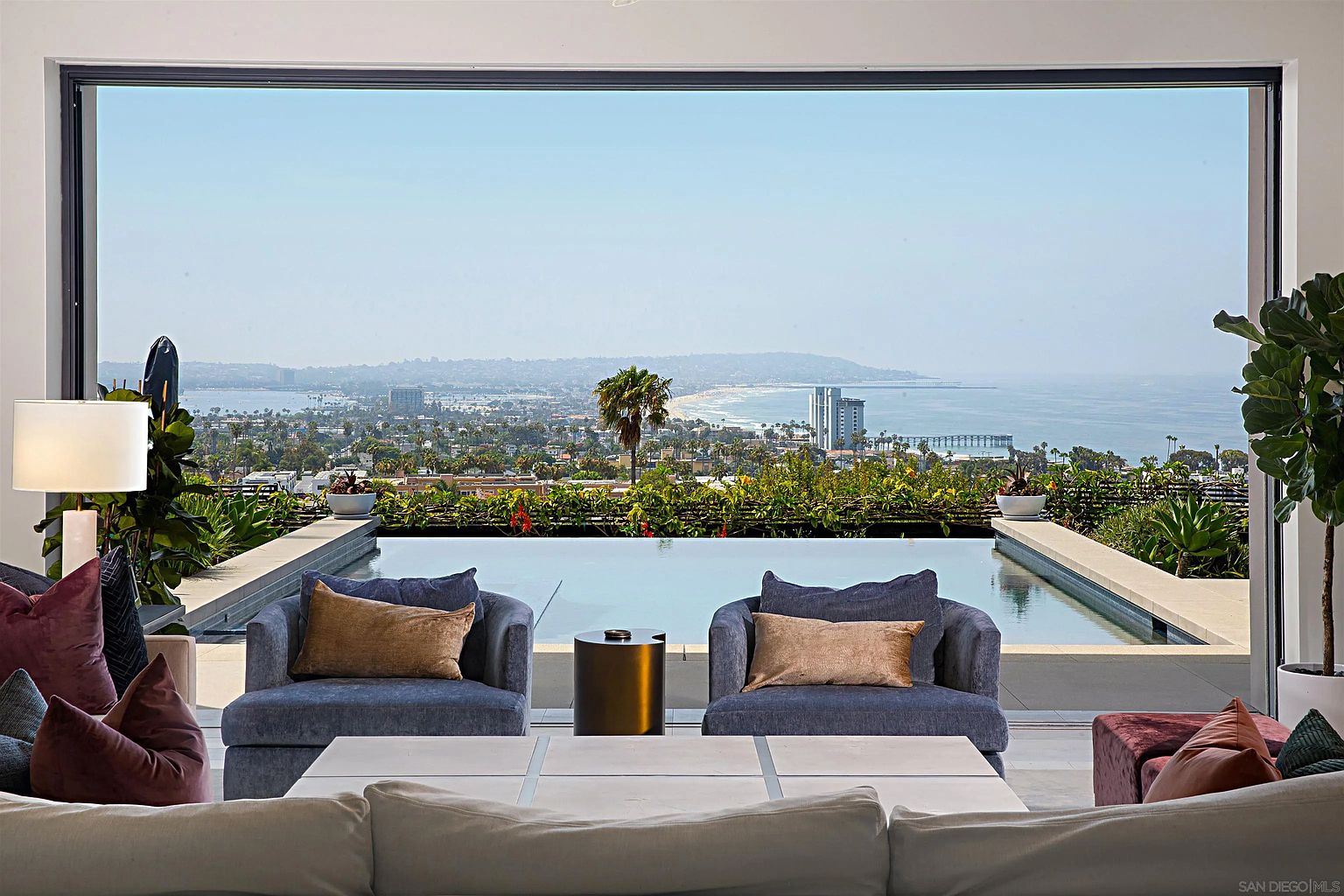 This image showcases a luxurious living room with a stunning view of the city and ocean through a large window. The room is furnished with comfortable armchairs, a modern coffee table, and plush pillows, creating an inviting and sophisticated atmosphere. An infinity pool is visible just outside the window, blending seamlessly with the horizon.