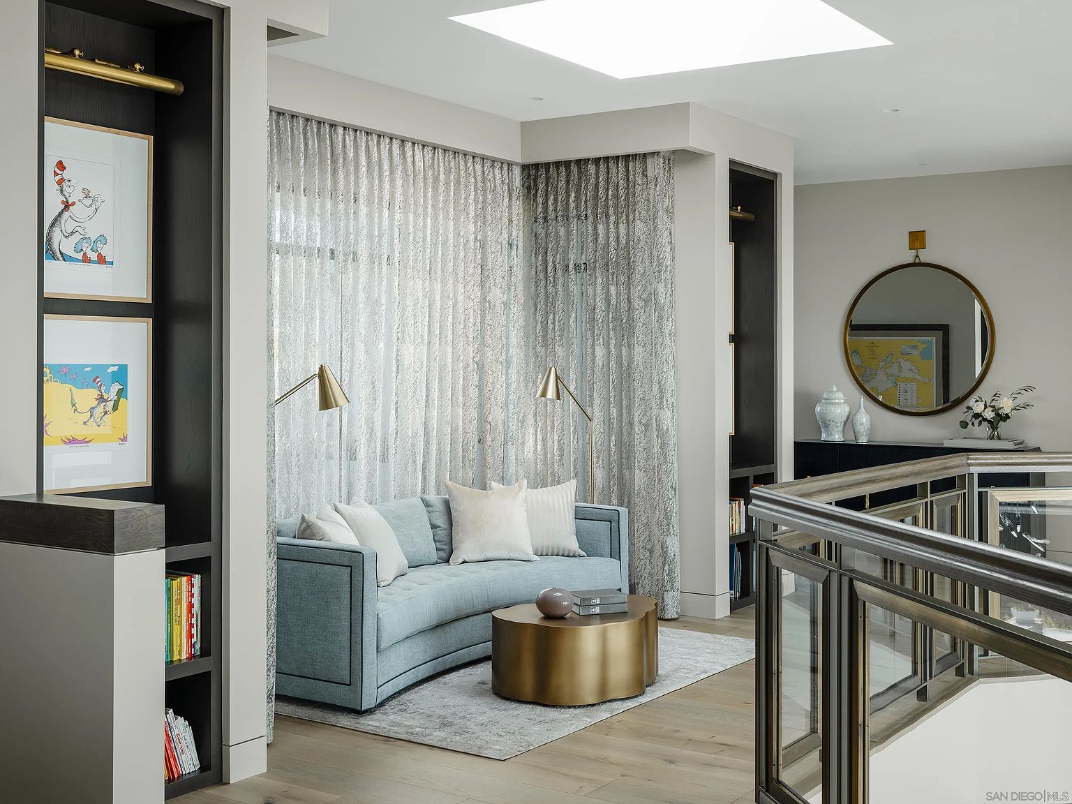 This interior shot showcases a stylish living room area with a curved blue sofa, a round gold coffee table, and elegant silver curtains. The room is well-lit with a skylight and features built-in shelving with artwork and books. The overall impression is modern and sophisticated, with a focus on comfort and design.