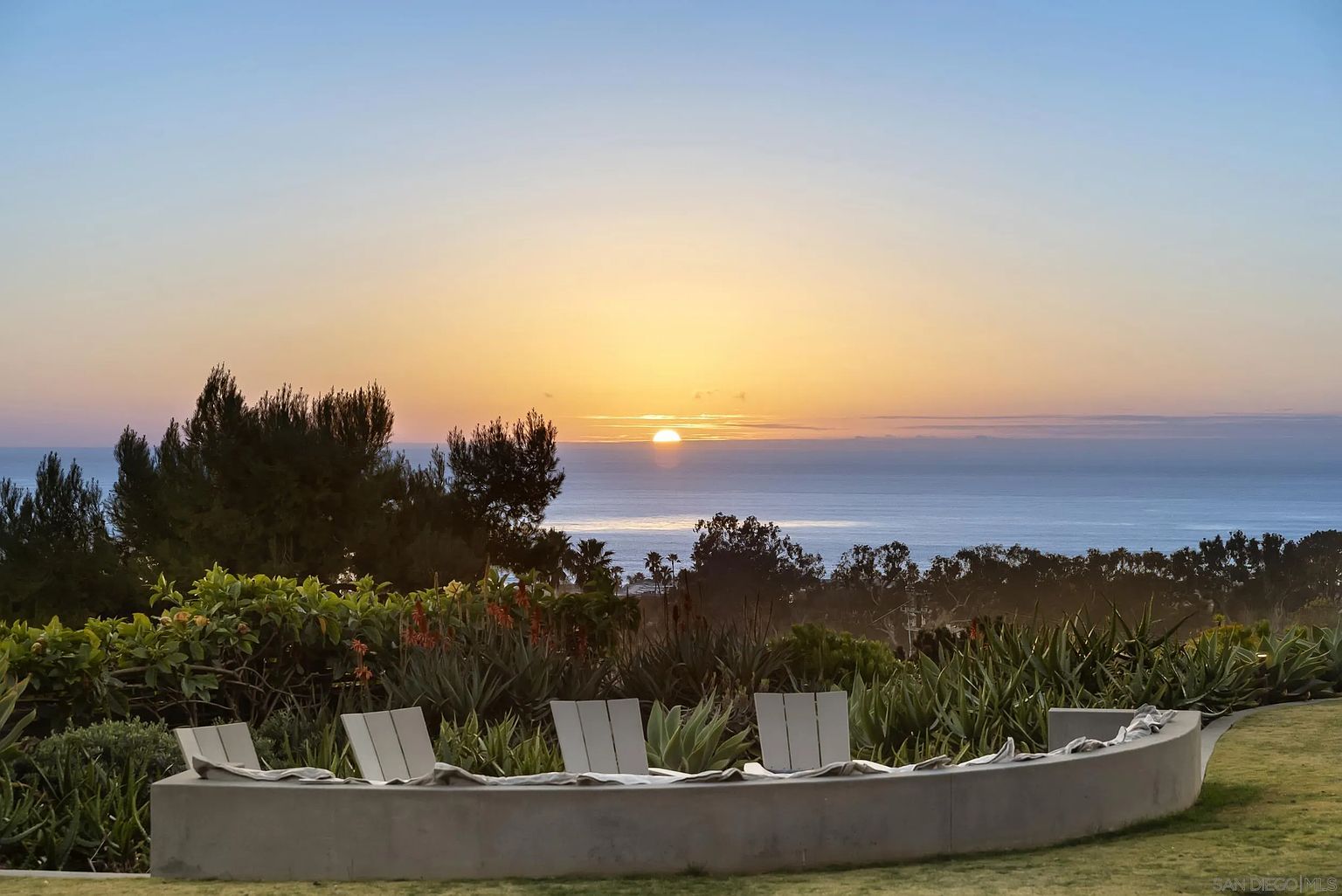 This image showcases a serene outdoor space with a stunning ocean view at sunset. A curved concrete seating area with cushions is surrounded by lush greenery, including various plants and trees. The scene evokes a sense of tranquility and relaxation, perfect for enjoying the natural beauty of the coastal landscape.