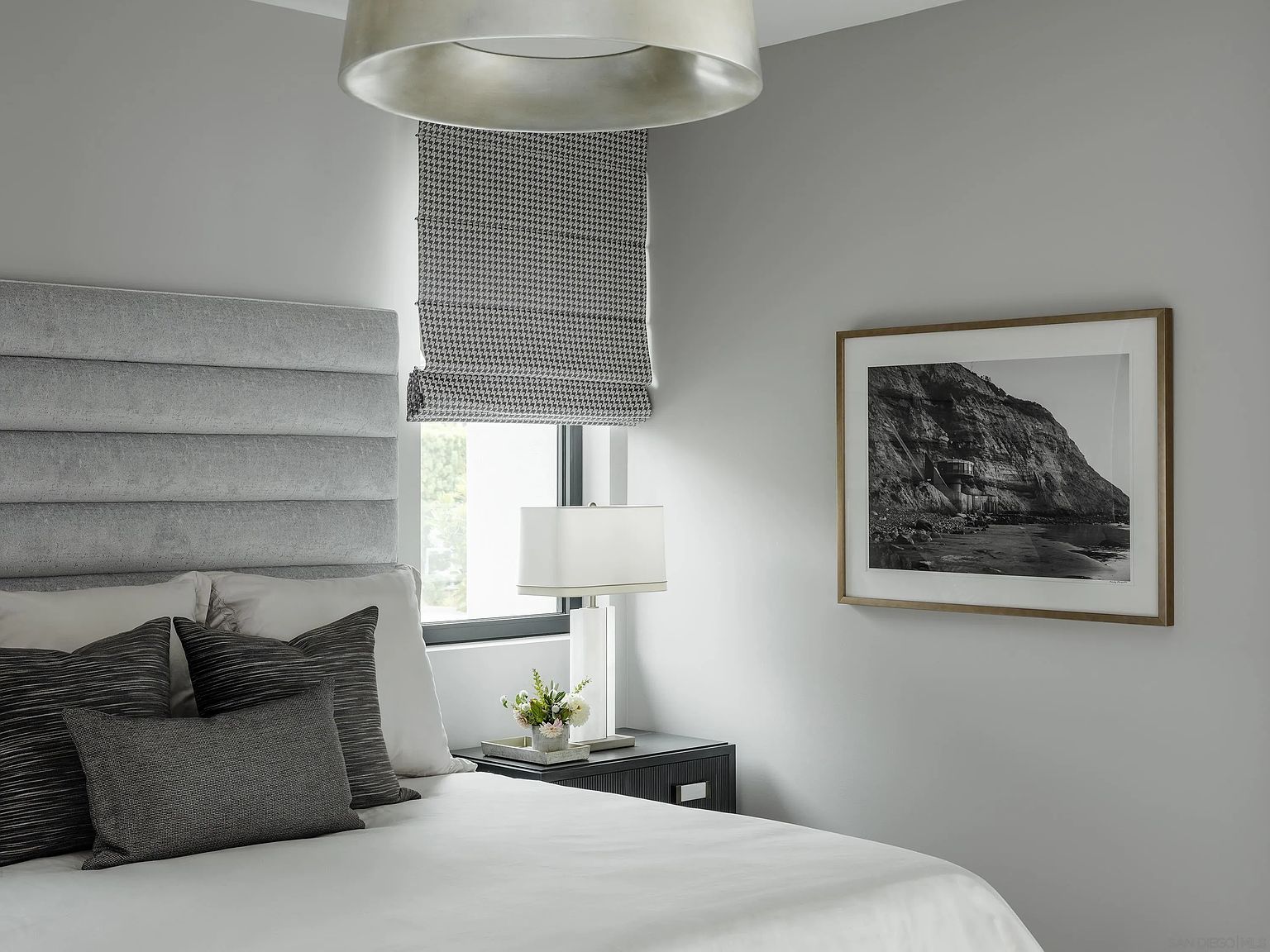 This is a well-lit primary bedroom featuring a large upholstered headboard, neutral color palette, and modern decor. A black and white photograph hangs on the wall, and a bedside table with a lamp and floral arrangement adds a touch of elegance. The room has a calm and sophisticated atmosphere.