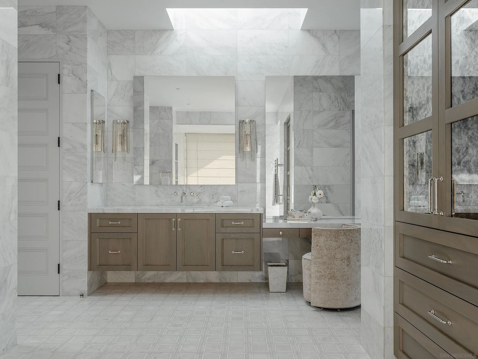 This is a luxurious primary bathroom featuring marble walls and flooring with a geometric pattern. A double vanity with a light wood finish is centered beneath two large mirrors, each flanked by sconces. A makeup vanity with a cushioned stool is to the right, and a tall linen cabinet is on the right side of the frame. The room is well-lit with natural light from a skylight, creating a bright and airy atmosphere.