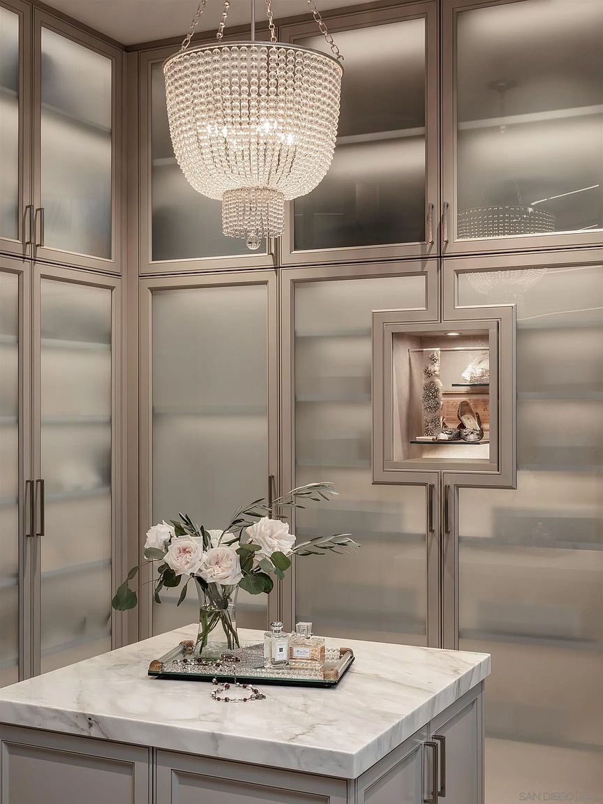 This image showcases a luxurious walk-in closet with frosted glass doors and a stunning crystal chandelier. A marble-topped island displays a vase of white roses and perfume bottles, adding a touch of elegance. The closet's design emphasizes organization and sophistication, creating a high-end, personalized space.