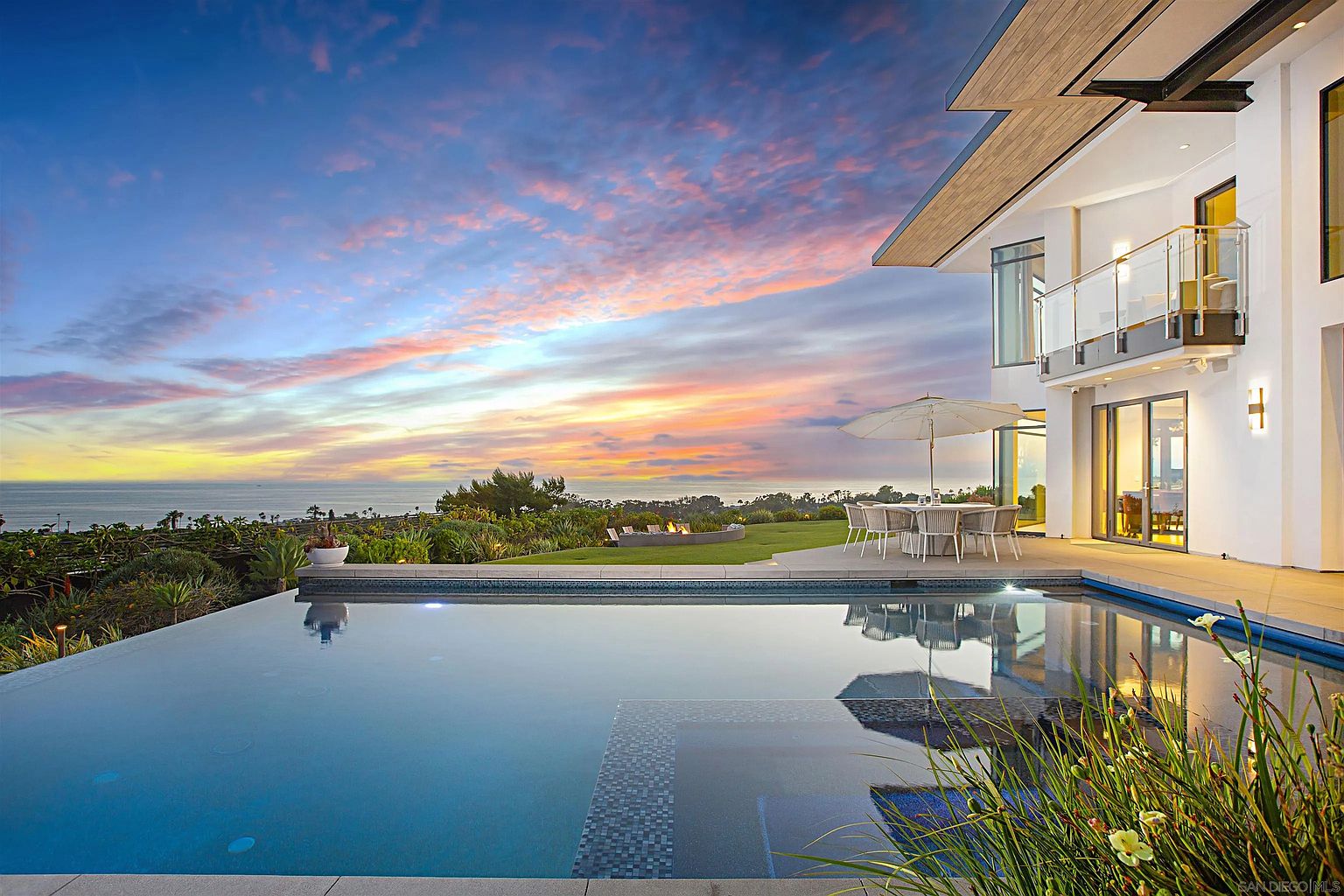 This image showcases a luxurious outdoor space featuring a modern home with a stunning infinity pool overlooking the ocean at sunset. The pool's design seamlessly integrates with the surrounding landscape, which includes lush greenery and a dining area with an umbrella. The overall impression is one of high-end living and serene relaxation.