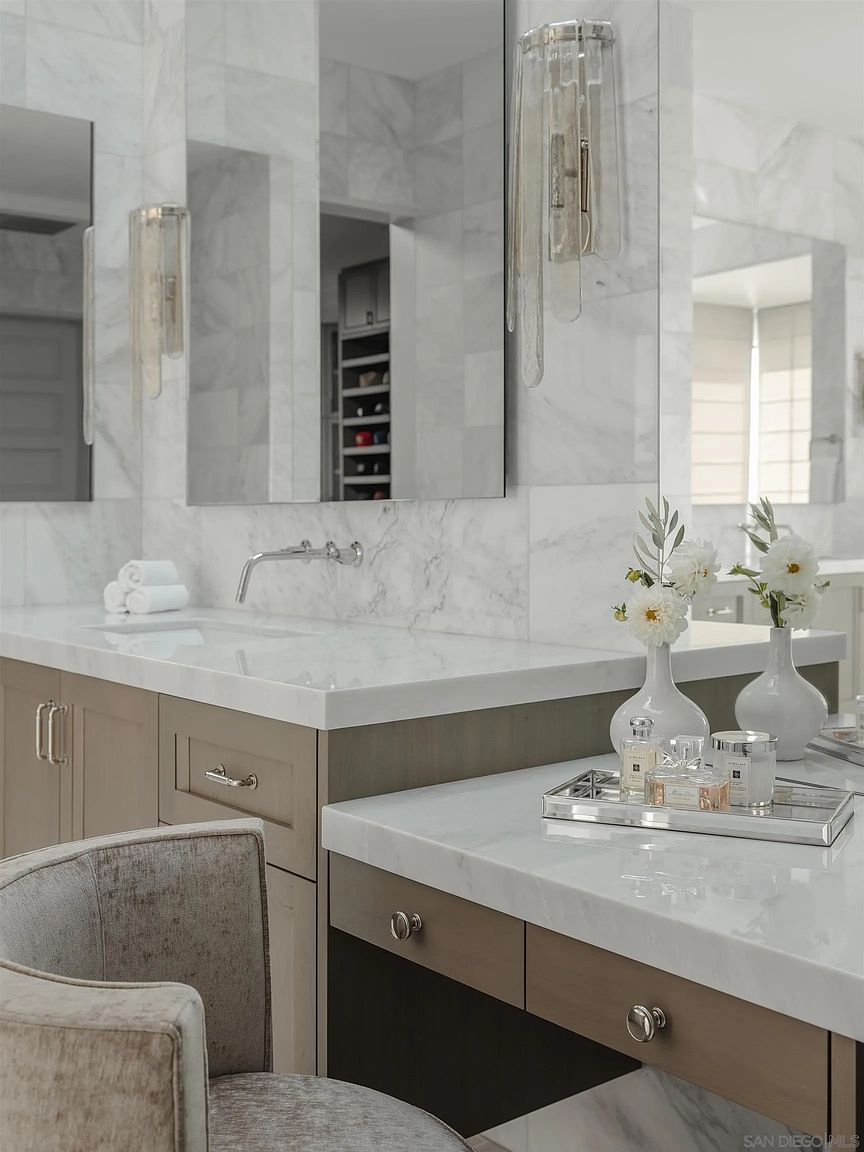 This is a luxurious primary bathroom featuring marble walls and countertops, creating a bright and elegant space. The vanity area includes a sink, drawers, and a dedicated makeup area with decorative items like perfume bottles and flowers. The overall impression is clean, modern, and upscale, perfect for a high-end real estate listing.