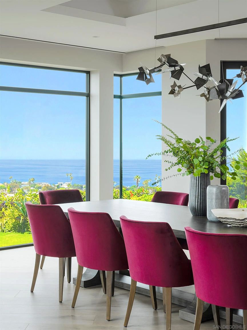 This is an interior shot of a dining room featuring a large table with seating for eight. The chairs are upholstered in a vibrant magenta fabric, adding a pop of color to the space. Large windows offer stunning views of the ocean and lush greenery, creating a bright and inviting atmosphere. A modern chandelier hangs above the table, adding a touch of elegance.