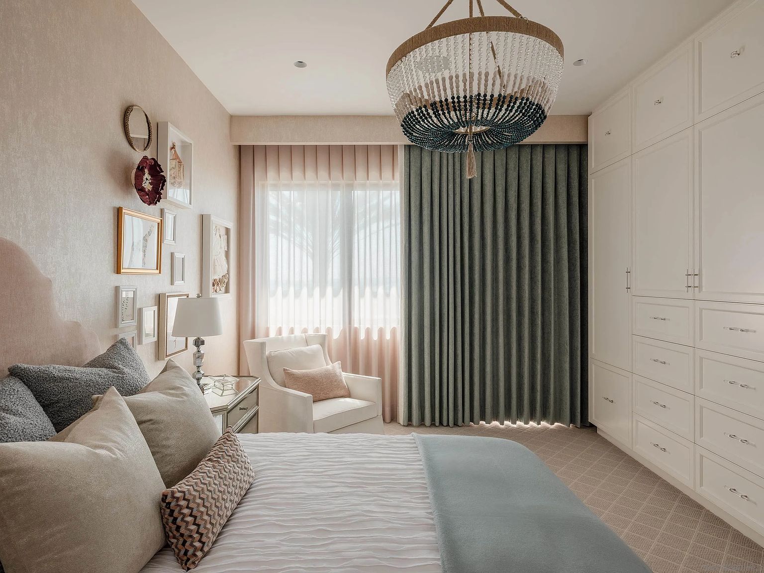 This is a serene primary bedroom featuring a plush bed with multiple pillows and a light blue throw. The room is decorated with a gallery wall of framed art and a stylish chandelier. The space is brightened by natural light filtering through sheer and blackout curtains, complemented by a large white wardrobe, creating a calm and inviting atmosphere.